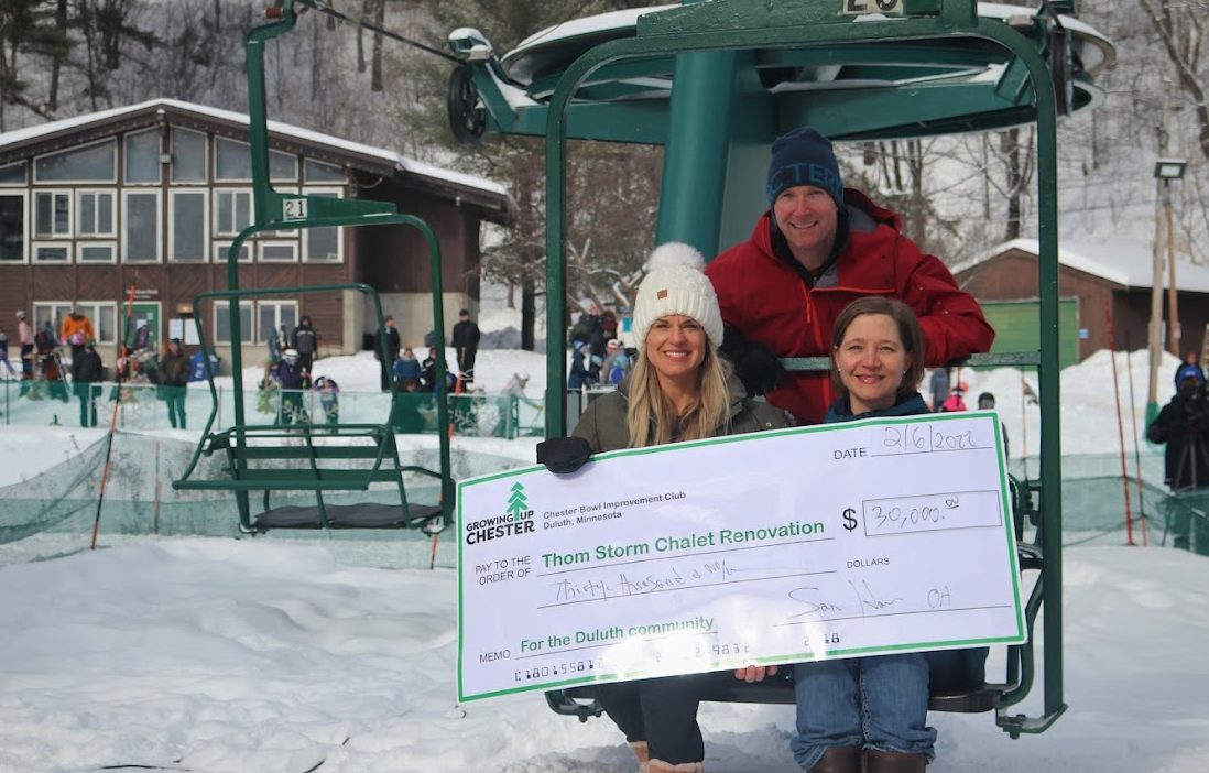 3 people sit on a ski lift chair holding a large check